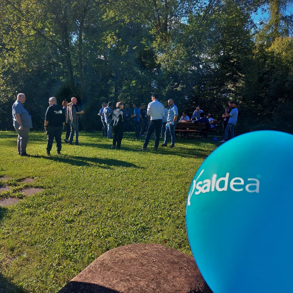 Palloncino blu con logo Saidea in evidenza, dietro un prato verde con i collaboratori di Saidea intenti a chiacchierare