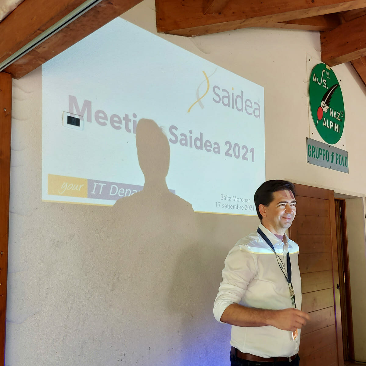 Al di fuori di una baita di montagna, il CEO di Saidea William Nicolussi presenta illuminato dalla slide proiettata sul muro con il testo "Meeting Saidea 2021"