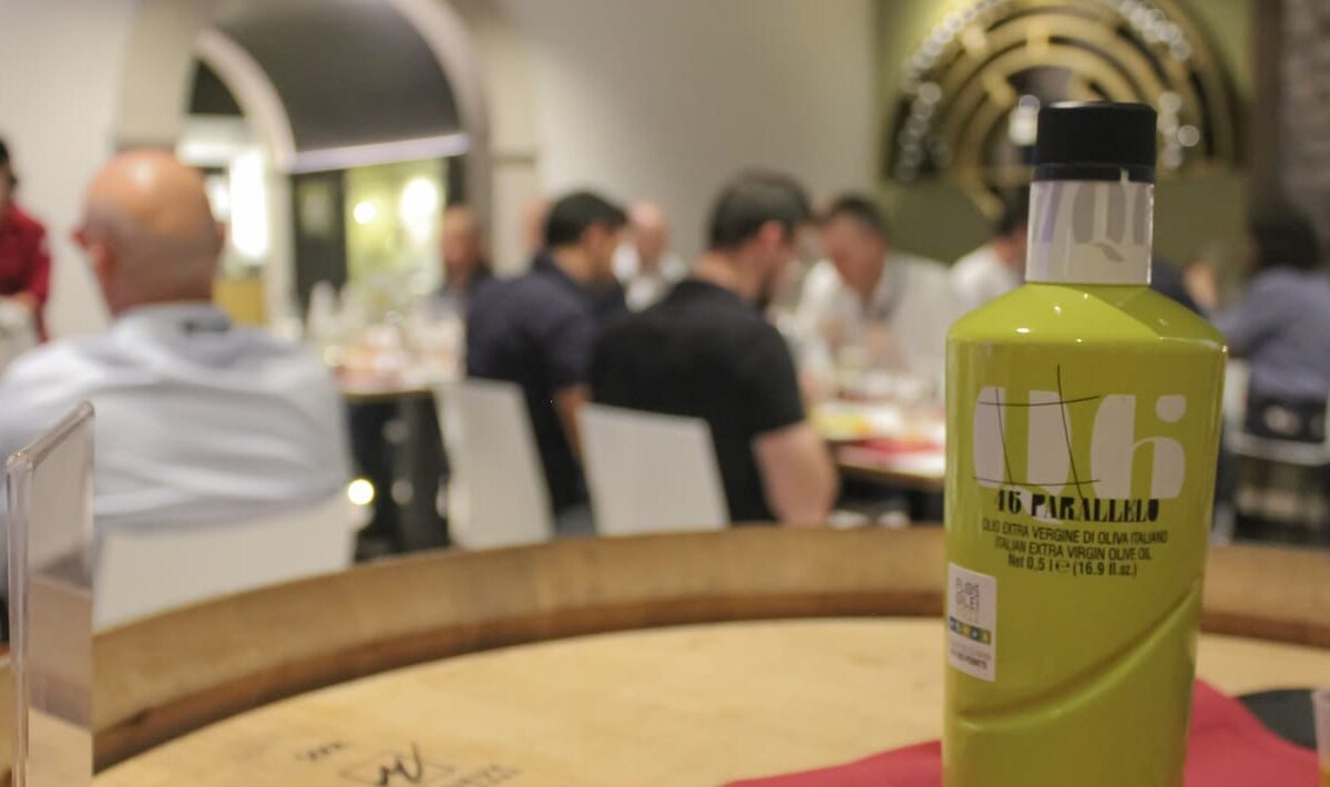 Una bottiglia di olio in primo piano, dietro ospiti, panelist e organizzatori degustano vini e oli del frantoio-cantina Agraria di Riva del Garda